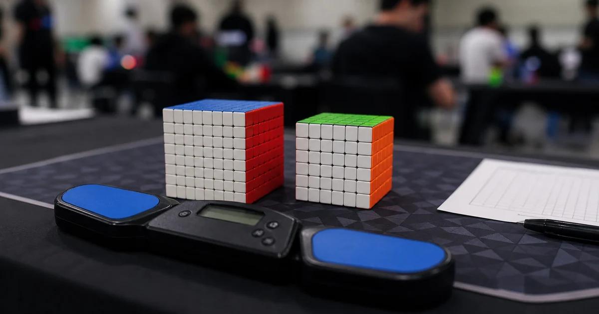 A 7x7 cube on a speedcubing mat next to a Stackmat timer and score sheet on a competition table, with a blurred venue background.