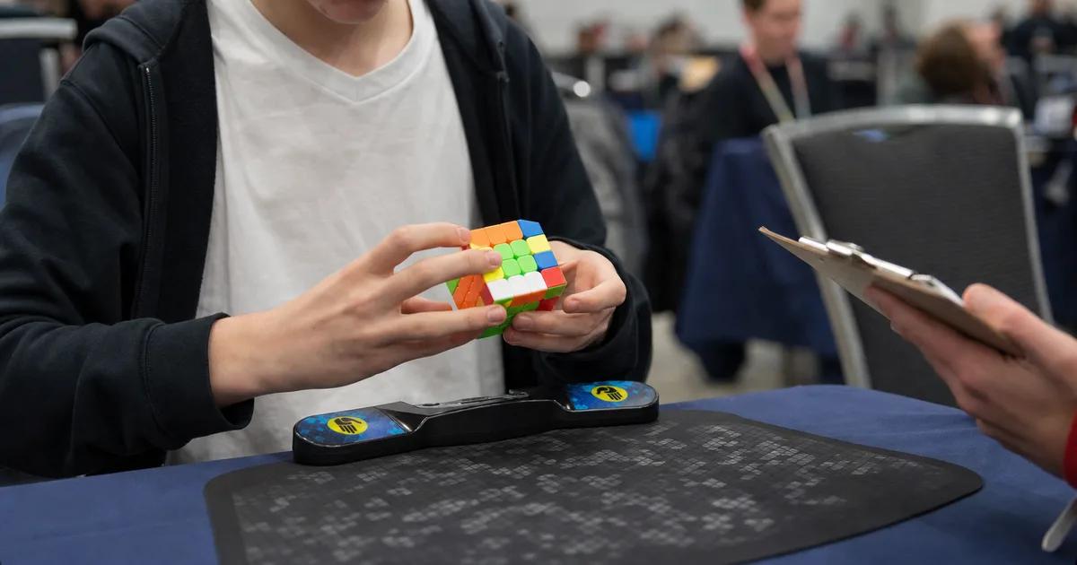A speedcuber solving a 3×3 cube on a Stackmat timer at a competition table, with score sheets and a judge in the background.
